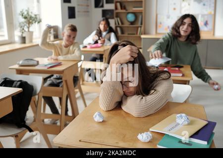 Offeso scolaretta seduta dalla sua scrivania, mentre un gruppo di compagni di classe crudeli gettando carte stropicciate a lei e schernendo a lei Foto Stock