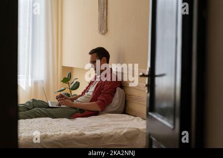Giovane uomo caucasico concentrato lavora come giornalista internet digitando articolo si siede sul letto di casa Foto Stock