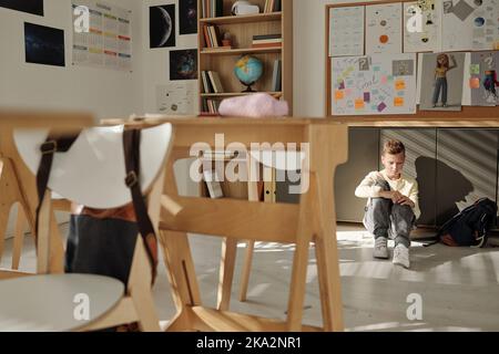 Scolaro solitario e infelice seduto sul pavimento di una classe vuota dopo le lezioni, mentre soffriva di bullismo dei compagni di classe Foto Stock