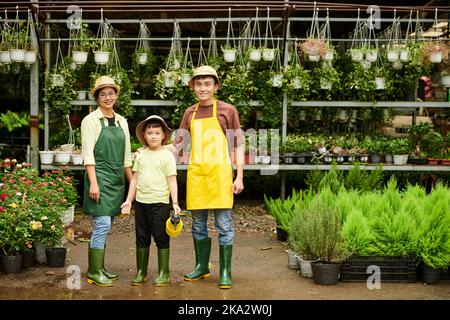 Genitori e figlio felici in stivali di gomma e grembiuli in piedi nel loro negozio di fiori Foto Stock