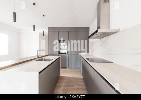 Cucina con lunghi ripiani in marmo bianco con una parete coperta da mobili di design grigio liscio e una colonna di elettrodomestici Foto Stock