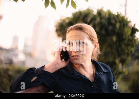 Donna d'affari matura che parla sullo smartphone seduto sul panca in città Foto Stock