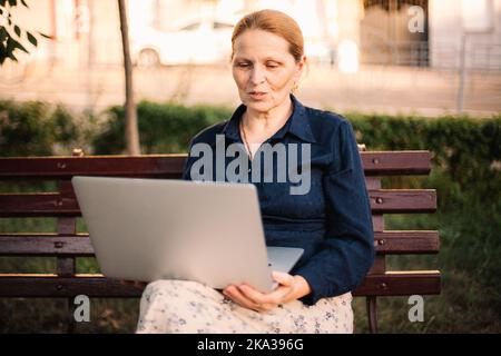 Donna matura che utilizza un computer portatile con videochiamata seduta sulla panca Foto Stock