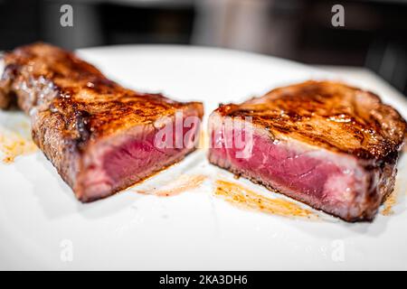 Macro primo piano di rara bistecca di manzo di carne alla griglia rossa cotta e alimentata con erba di New York tagliata in due metà con crudo all'interno su piatto bianco Foto Stock