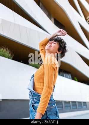 Da sotto vista laterale di Happy Hispanic femmina con capelli corti ricci in denim generale in piedi con gli occhi chiusi in città strada vicino creativo buil urbano Foto Stock