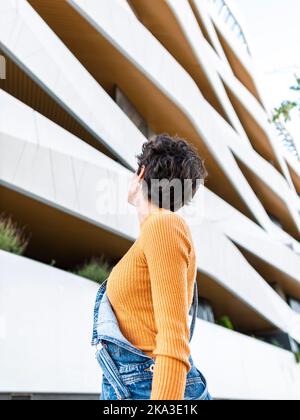 Dal basso, vista posteriore di una donna irriconoscibile con capelli ricci corti in denim, in generale, che si erge sulla strada cittadina e che ammira il creativo edificio urbano Foto Stock