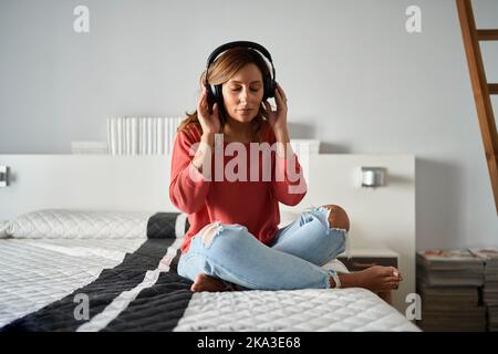Donna ispanica di mezza età in abiti casual che toccano le cuffie e chiudono gli occhi mentre si siede a gambe incrociate sul letto e l'ascolto di musica nel fine settimana Foto Stock