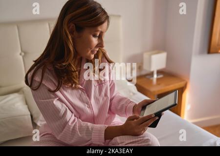 Donna matura in pajama rosa seduta a letto e lettura e libro nel fine settimana di sole mattina a casa Foto Stock