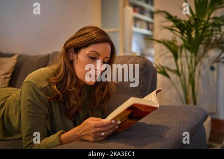 Vista laterale di una donna ispanica matura positiva sdraiata sul divano e libro di lettura nel fine settimana sera in soggiorno Foto Stock