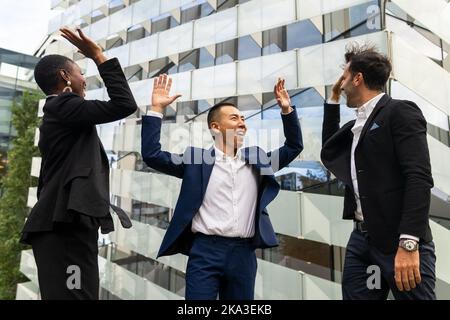 Felici colleghi multirazziali in abiti formali in piedi contro edificio urbano di vetro e dando alti cinque a vicenda, mentre celebrare il successo de Foto Stock