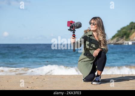 Giovane donna con capelli lunghi in casual trench coat seduta su fienili su spiaggia sabbiosa vicino mare ondeggiante e registrazione vlog tramite fotocamera professionale con mi Foto Stock