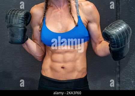 Sportswoman muscolare irriconoscibile in abbigliamento sportivo e guanti che mostrano i muscoli contro la parete grigia durante l'allenamento di pugilato in palestra Foto Stock