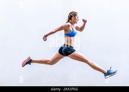 Atleta femminile muscolosa in corporatura piena determinata in braccio di sollevamento di Active Wear e saltellando contro la parete bianca durante l'addestramento intenso di idoneità Foto Stock