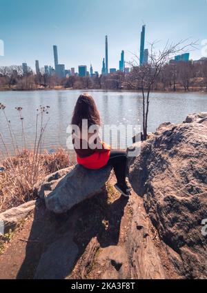 Corpo pieno femmina irriconoscibile con lunghi capelli seduti su masso e ammirando le calme acque dello stagno di Central Park nel giorno di autunno nuvoloso a New York, Foto Stock