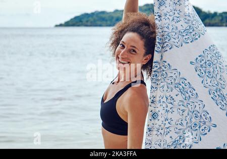 Vista laterale di Happy FIT donna ispanica con capelli ricci tenendo leggero telo da spiaggia in piedi vento sulla costa e guardando una macchina fotografica Foto Stock