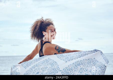 Vista posteriore di Happy FIT donna ispanica con capelli ricci tenendo leggero panno da spiaggia in piedi vento sulla costa e guardando una macchina fotografica Foto Stock