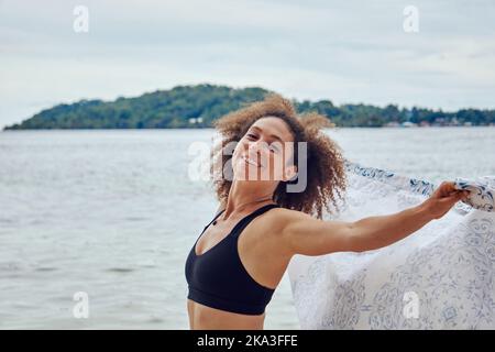 Vista laterale di Happy FIT donna ispanica con capelli ricci tenendo leggero telo da spiaggia in piedi vento sulla costa e guardando una macchina fotografica Foto Stock