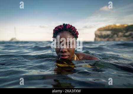 Giovane afroamericana che guarda la fotocamera mentre nuota in mare contro il cielo durante le vacanze estive Foto Stock