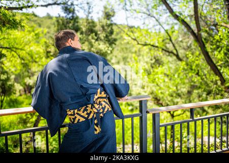 Giovane uomo in costume kimono abiti tradizionali in piedi con la schiena, appoggiato su ringhiera recinto nel giardino all'aperto in Giappone con vista sulla natura Foto Stock