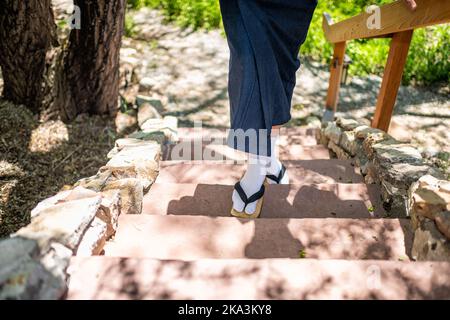 Piedi dell'uomo in costume kimono, calze di scarpe geta tabi in giardino esterno con scale in pietra in Giappone da ringhiera in legno Foto Stock