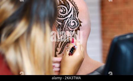 Una ragazza, padrone del tatuaggio, artista del mehendi fa disegno del tatuaggio dell'henné sul cuoio capelluto dell'uomo calvo caucasico, spalla, collo.il processo di applicazione dell'henné da un tubo alla pelle. Foto di alta qualità Foto Stock