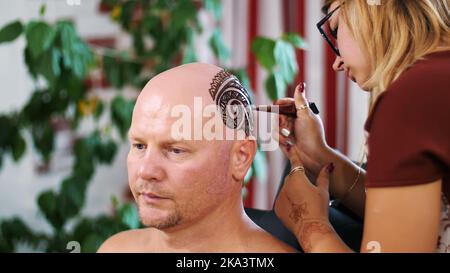 Una ragazza, padrone del tatuaggio, artista del mehendi fa disegno del tatuaggio dell'henné sul cuoio capelluto dell'uomo calvo caucasico, spalla, collo.la soluzione dell'henné asciuga sulla pelle. Foto di alta qualità Foto Stock