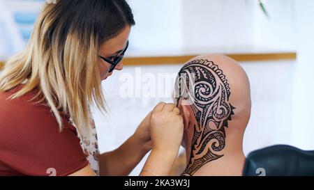 Una ragazza, padrone del tatuaggio, artista del mehendi fa disegno del tatuaggio dell'henné sul cuoio capelluto dell'uomo calvo caucasico, spalla, collo.il processo di applicazione dell'henné da un tubo alla pelle. Foto di alta qualità Foto Stock