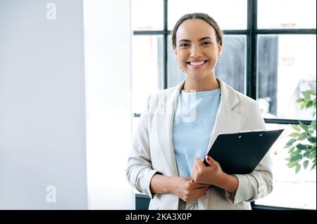 Foto di una donna d'affari positiva brasiliana o latina di successo in piedi in un ufficio di lavoro con una cartella di documenti nelle sue mani, manager finanziario, realtor, guardando la fotocamera, sorrisi Foto Stock