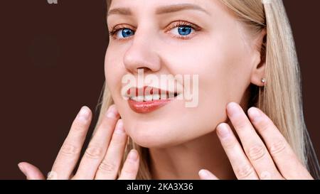 Faccia di bella donna sorridente con trucco naturale guardando la macchina fotografica primo piano. La giovane ragazza tocca il suo volto con una pelle chiara e brillante. Prolungamento di Foto Stock