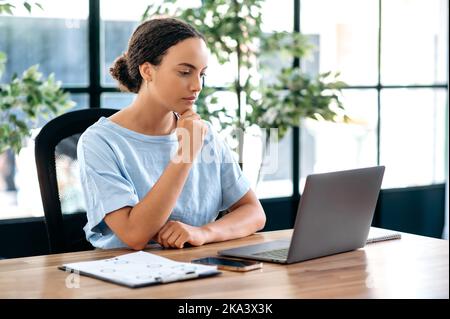 Foto di un latino o di una donna brasiliana, dipendente dell'ufficio, manager creativo, seo, si siede sul posto di lavoro in ufficio, guardando il portatile concentrato, pensando a un progetto, la strategia di pianificazione Foto Stock