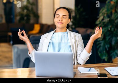 Meditazione, relax. Calma bella donna di affari di razza mista, collaboratore aziendale, responsabile del rischio, medita con gli occhi chiusi sul posto di lavoro, prende una pausa dal lavoro, curando la sua salute mentale Foto Stock