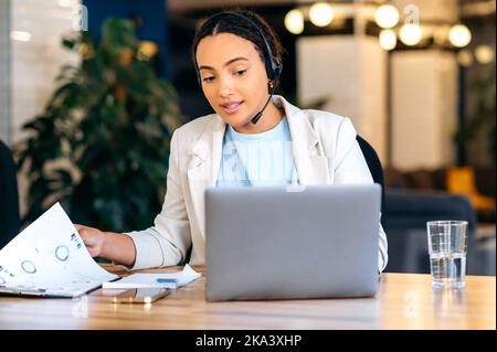 Donna d'affari brasiliana o ispanica affollata, consulente, operatore del call center, con cuffie, seduto alla scrivania in ufficio, consultando in remoto il cliente, fornendo consigli, rispondendo alle domande Foto Stock