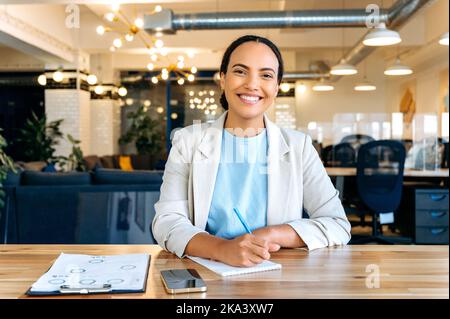 Bella giovane brasiliana o latino donna di successo, mentore d'affari, consulente finanziario, studente, guardando la webcam durante la consultazione o webinar da videoconferenza, prende appunti in notebook, sorridendo Foto Stock