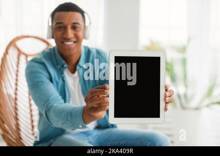 Felice giovane uomo afroamericano con cuffie wireless che mostrano un tablet con schermo vuoto Foto Stock
