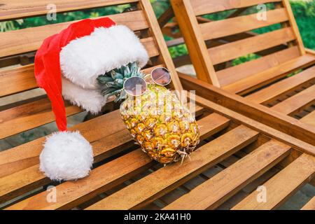 Vista laterale del rilassante e divertente ananas indossando occhiali da sole e il cappello di Babbo Natale si trova sul lettino in legno durante il sole nei tropici. Natale ai tropici Foto Stock
