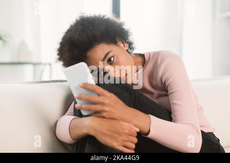 Triste depressa giovane afroamericana con capelli ricci abbracciando le ginocchia e guardando smartphone Foto Stock