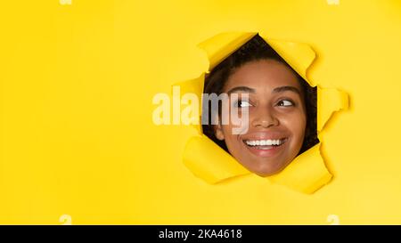 Donna nera che guarda da parte attraverso il foro nella carta, sfondo giallo Foto Stock