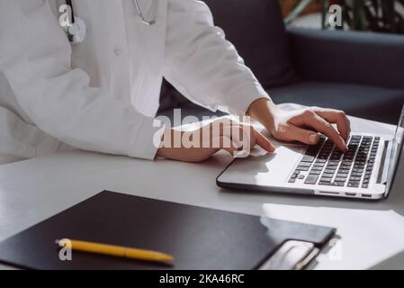 Primo piano delle mani di una dottoressa che digita sulla tastiera di un computer portatile seduto su un comodo divano alla scrivania dell'ufficio della clinica. Donna infermiera con uniforme bianca a Foto Stock