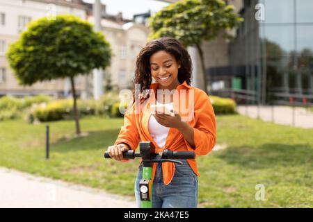 Felice giovane afro-americana femmina con scooter elettrico a digitare sullo smartphone, divertirsi in estate Foto Stock