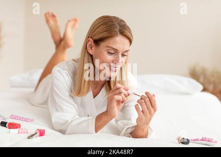 Donna matura felice applicando smalto del chiodo, facendo manicure, sdraiato sul letto, facendo la procedura domestica della stazione termale, copia lo spazio Foto Stock