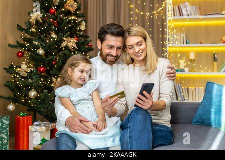 Famiglia felice per lo shopping di Natale in linea che sceglie i regali nel deposito in linea, uomo e donna con il bambino che siede nel paese sul divano vicino all'albero di Natale che tiene la carta di credito della banca. Foto Stock