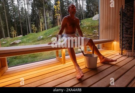 Rilassante uomo avvolto bianco asciugamano seduto sulla panca di legno nella sauna finlandese calda con una grande finestra con vista verde della foresta e godere piacevole Foto Stock