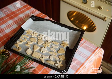 Vista dall'alto foglio da forno con stampi di pasta di pan di zenzero tagliati sul tavolo da cucina sullo sfondo di un forno di riscaldamento Foto Stock