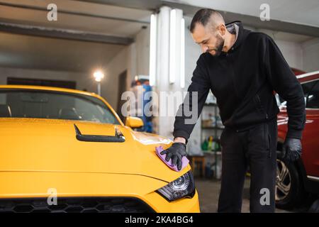 Dettaglio e concetto di pulizia dell'auto. L'operaio del lavaggio dell'automobile maschio tiene la microfibra in mano e lucida il cofano dell'automobile. Messa a fuoco selettiva sulla mano con microfibra. Foto Stock