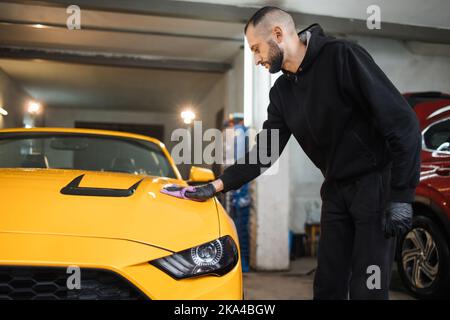 Dettaglio e concetto di pulizia dell'auto. L'operaio del lavaggio dell'automobile maschio tiene la microfibra in mano e lucida il cofano dell'automobile. Messa a fuoco selettiva sulla mano con microfibra. Foto Stock