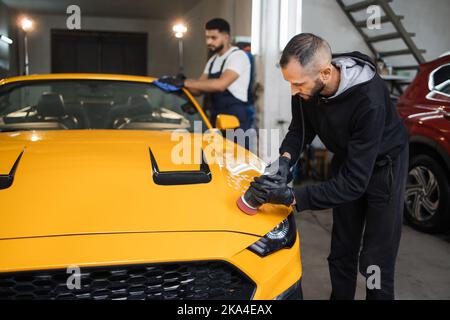 Team di giovani uomini bearded, lavoratori di auto servizio di dettaglio. I maschi sono in grado di tenere in mano la lucidatrice orbitale e lucidare il cappuccio e pulire il parabrezza dell'auto moderna gialla. Concetto di dettaglio e lucidatura dell'auto. Foto Stock