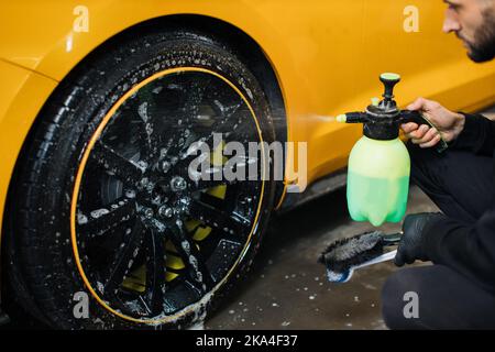 Pulizia cerchi auto, concetto di lavaggio dettagli auto. Foto ravvicinata di mano maschile in guanto di gomma nera con spruzzatore, lavaggio ruota in lega per auto al lavaggio auto e servizio di dettaglio. Foto Stock