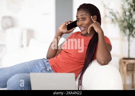 Giovane donna nera che parla sul cellulare e lavora con il portatile a casa Foto Stock
