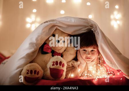 piccola ragazza sorridente carina con orsacchiotto bianco che sbircia fuori da sotto una coperta su un letto a casa con ghirlande illuminanti al buio. Bambino che guarda dentro Foto Stock