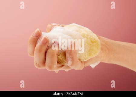 Un primo piano di una donna che strizza una spugna di mare gialla su uno sfondo di colore rosa Foto Stock